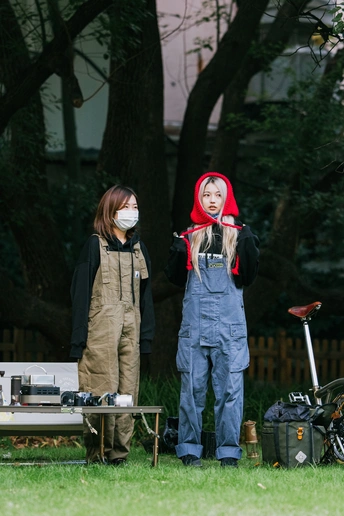 一个戴红面具的男子站在一个穿工作服的年轻女孩旁边 背景是一个有摩托车的院子。