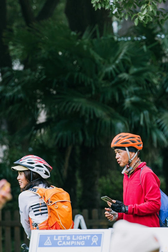 一名戴橙色头盔的自行车手正在看手机 而另一名骑自行车的人正在看着他。