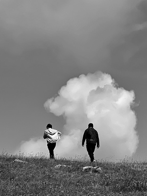 一张黑白照片 一对男子走在草地上 朝着充满云朵的天空走去。