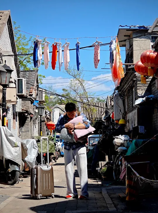 一位女士抱着一个婴儿 带着一个手提箱 走过一个挂满晾衣绳的狭窄小巷。