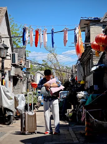 一位女士抱着一个婴儿 带着一个手提箱 走过一个挂满晾衣绳的狭窄小巷。