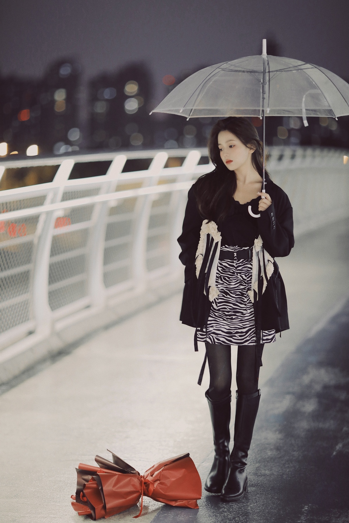 一位穿着黑白裙子的年轻女子站在一座桥上 手里拿着一把伞 肩上背着一只红色的包。
