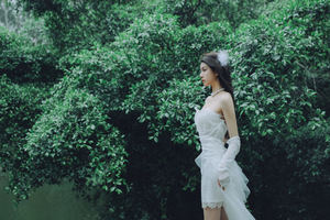 一位穿着白色连衣裙 拖尾的美丽年轻女子站在森林中 树木和水 手里拿着雨伞。