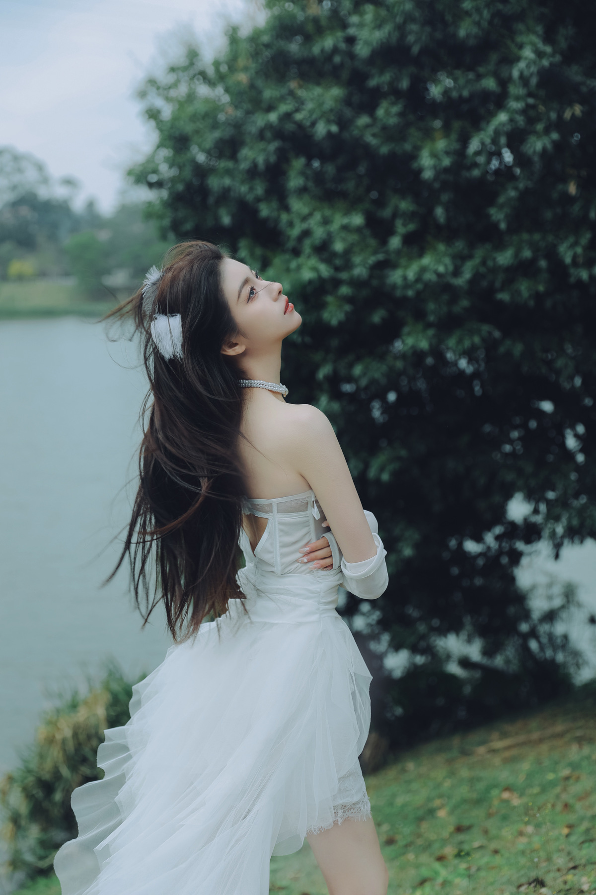 一位穿着白色连衣裙的年轻女子站在湖边草地上 背景中有树木和水。