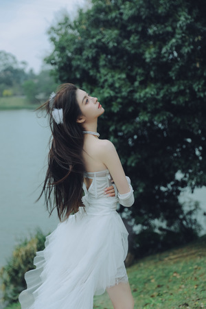一位穿着白色连衣裙的年轻女子站在湖边草地上 背景中有树木和水。