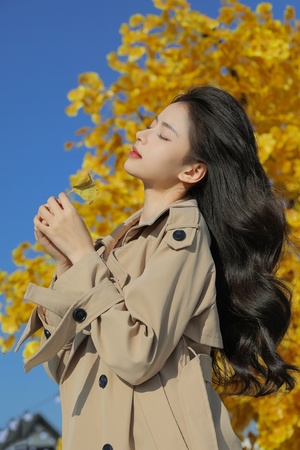 一位年轻女子穿着一件军大衣 手里拿着一枚黄色的树叶 抬头望向天空。