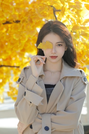 一位穿着黄色外套 脸上带着黄色树叶的年轻女子 站在黄色的树前 秋天。
