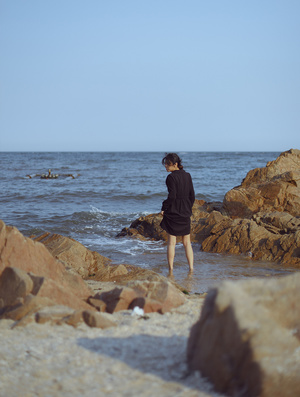 一个穿着黑色连衣裙的年轻女子站在靠近海洋的岩石海岸上 另一个人在水中走动。