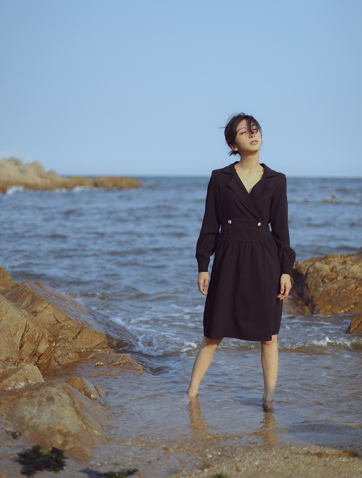 一位穿着黑色外套和连衣裙的女人在靠近海洋的岩石海滩上行走。