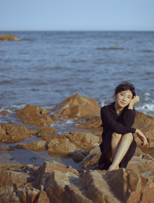 一个穿着黑色连衣裙的年轻女子坐在海洋边一块岩石上
