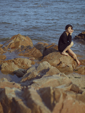 一位穿着连衣裙的年轻女子坐在靠近海洋的岩石海岸上的一些岩石上。
