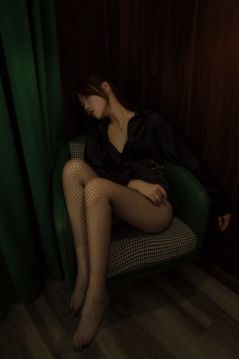 一个穿着黑色连衣裙和长筒袜的年轻女子坐在一个绿色椅子上 在一个黑暗的房间里。