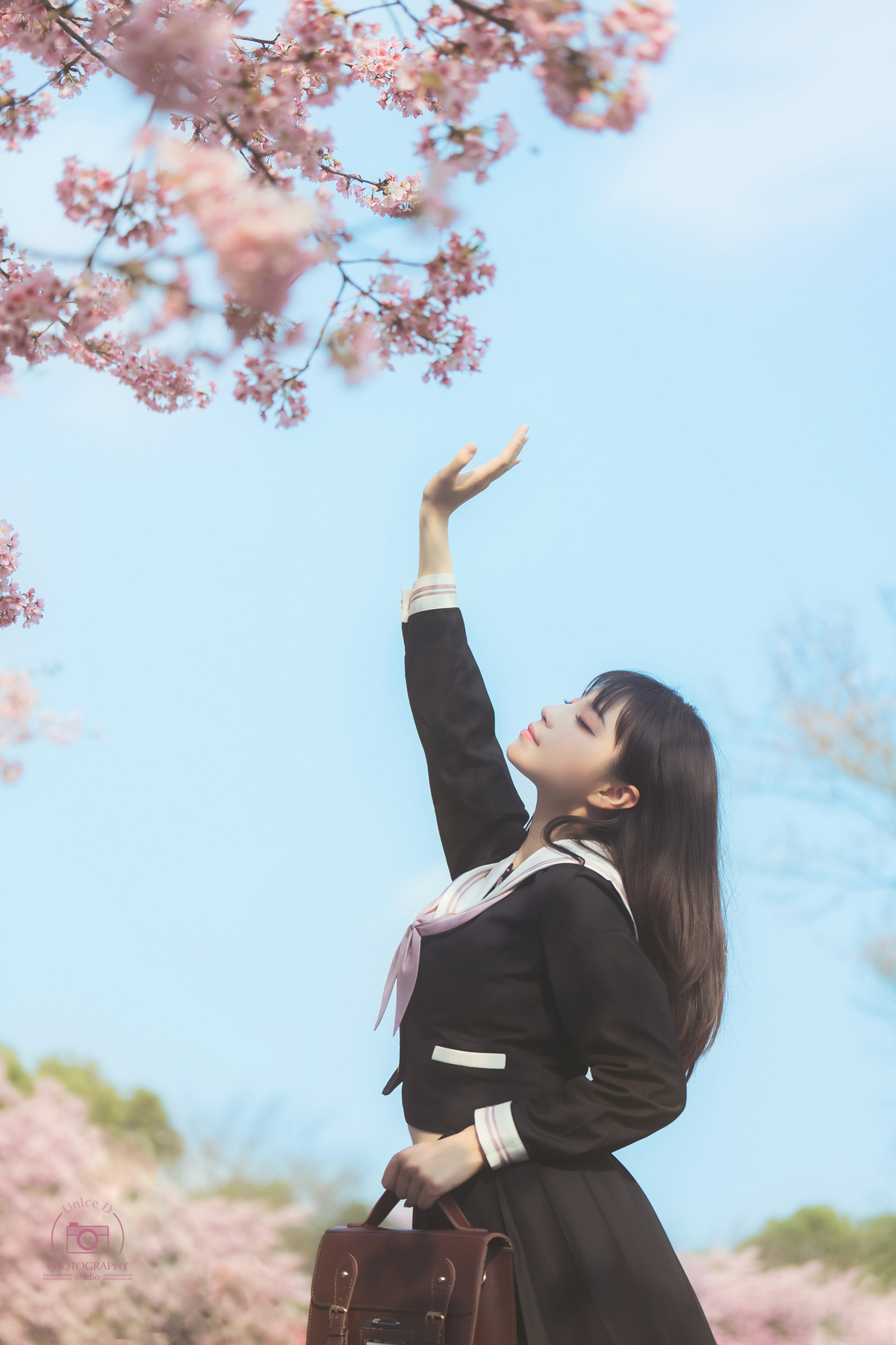 一个年轻的女人站在一棵开着粉红色花朵的树前 手里拿着一个手提箱 抬头仰望天空