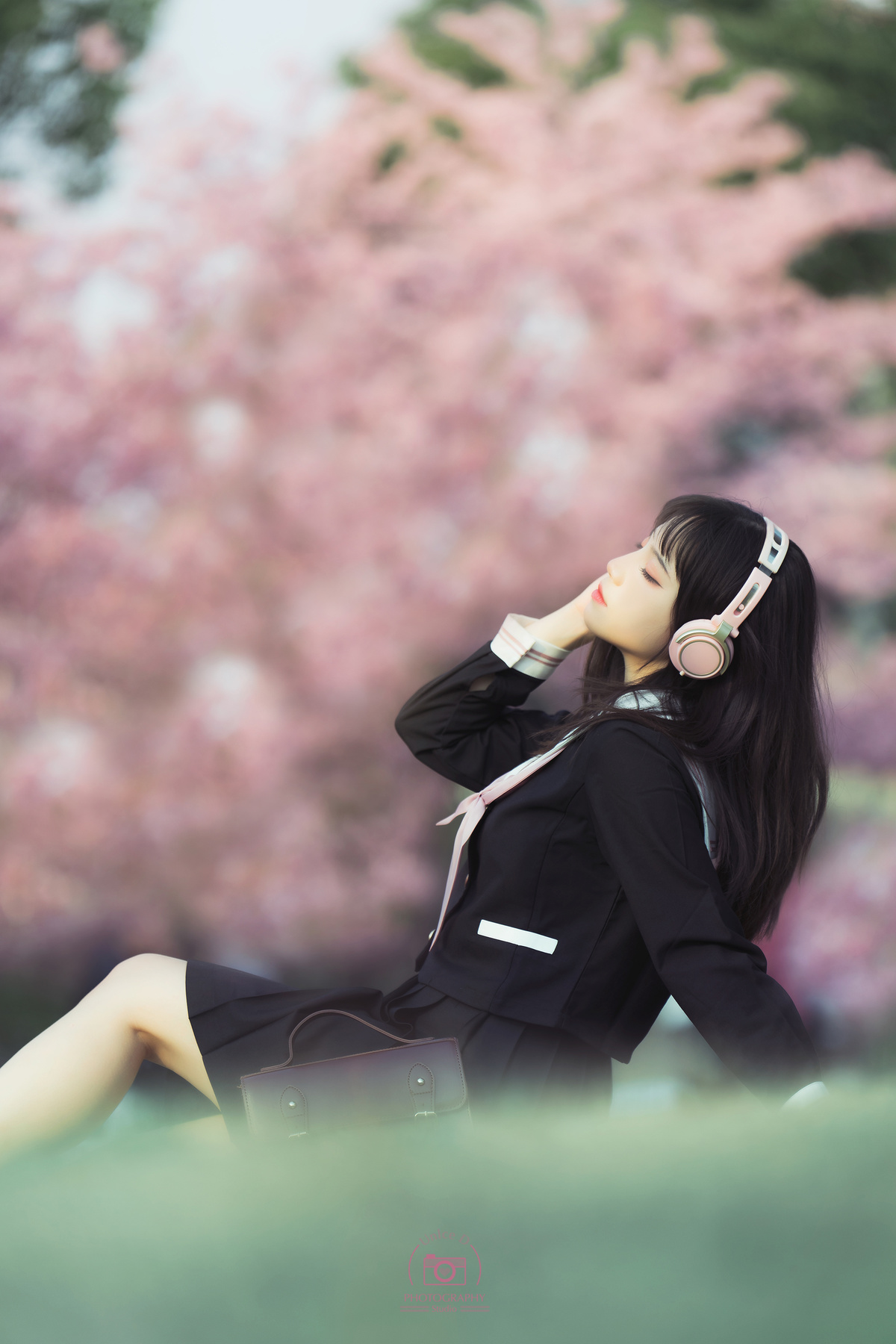 一位戴着耳机的年轻女子坐在一棵开着粉色花朵的树前的草地上 一边用手机听音乐