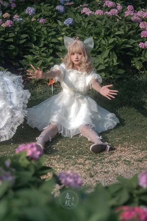 一个小女孩穿着天使装 带着白色翅膀 坐在花园前的草地上 身旁有一些花。