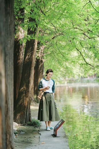 一位年轻女子站在一棵树旁的水边读书
