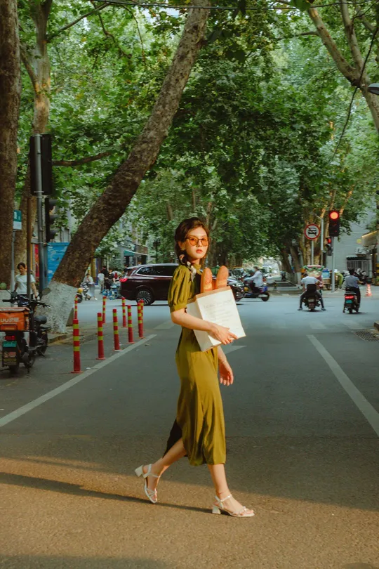 一位穿着绿色连衣裙的年轻女子拿着一张纸在一条栽满树街道上走下来