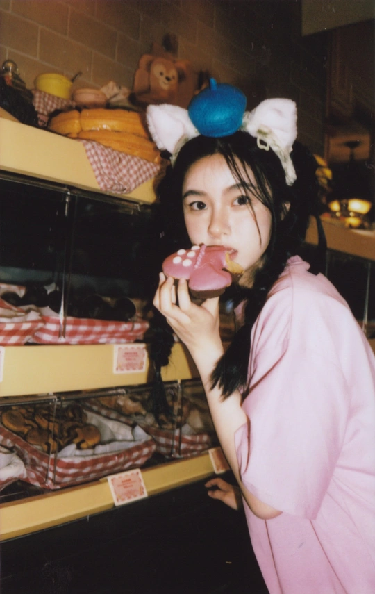 一位年轻女子咬着一个粉红色的甜甜圈站在一家商店的货架前。