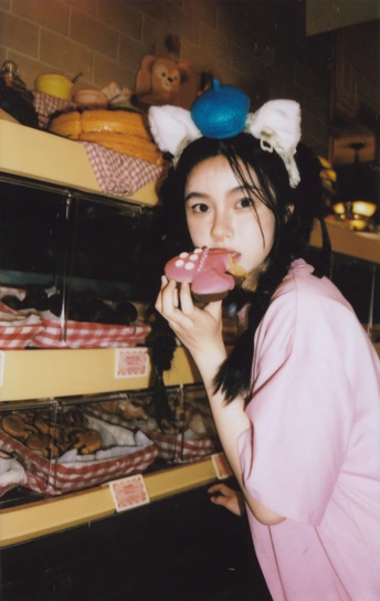 一位年轻女子咬着一个粉红色的甜甜圈站在一家商店的货架前。