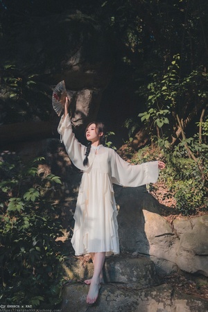 一位穿着白色长袍的年轻女子站在一些石头上 手里拿着一把扇子。