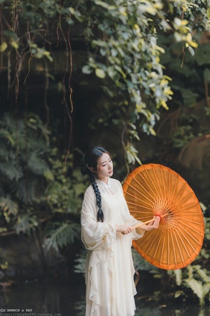 一位穿着白色连衣裙 手持橙色雨伞 站在茂密绿色森林小溪旁的女士