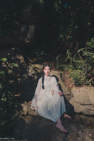 一位美丽的年轻女士穿着白色连衣裙坐在森林中的一块岩石上