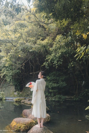 一位穿着白色连衣裙 手持雨伞站在流水中的岩石上的女人