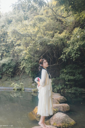 一位穿着白色连衣裙的年轻女子站在河中的一块岩石上 手里拿着一朵花。