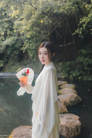 一位穿着传统服装的年轻女子手持扇子站在河边。