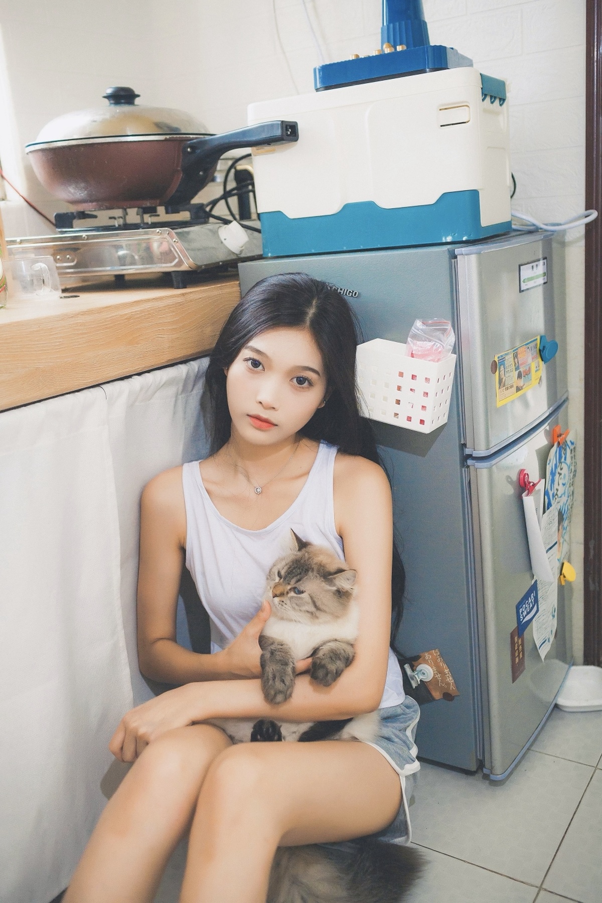 一位年轻女子抱着一只猫坐在冰箱前的购物袋上。
