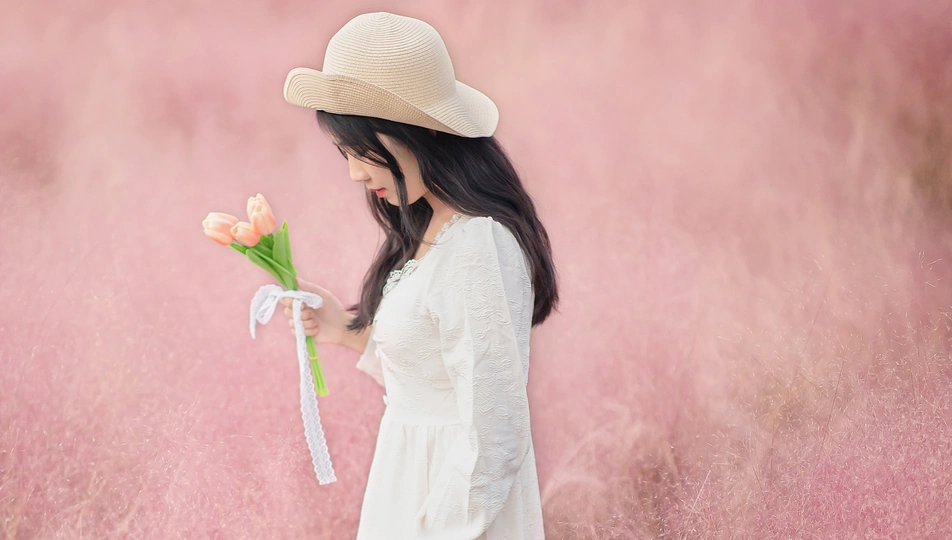 一位穿着白色连衣裙和帽子 手里拿着花站在粉红色田野上的美丽年轻女子。