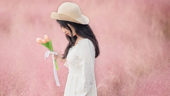 一位穿着白色连衣裙和帽子 手里拿着花站在粉红色田野上的美丽年轻女子。