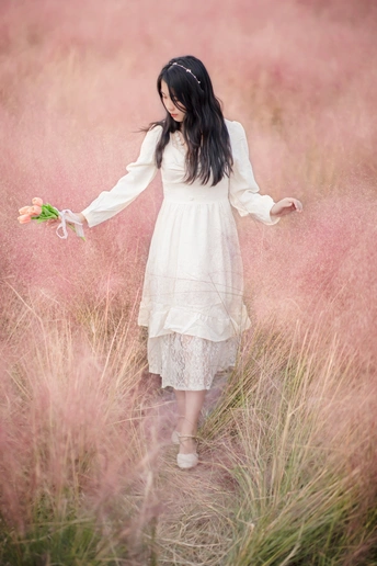 一个穿着白色连衣裙的年轻女子 手里拿着一朵花 站在一片高草地上。