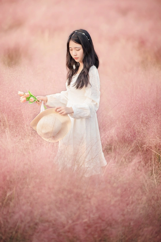 一位穿着白色连衣裙和帽子 手里拿着一朵花的年轻女子 站在一片高草地上。