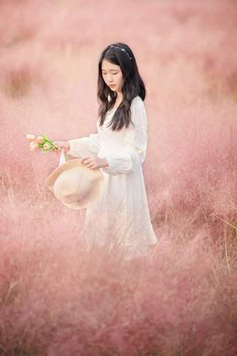 一位穿着白色连衣裙和帽子 手里拿着一朵花的年轻女子 站在一片高草地上。