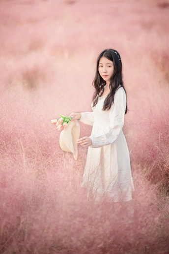 一位穿着白色连衣裙和帽子 手里拿着一朵花的年轻女子 站在粉红色草地上。