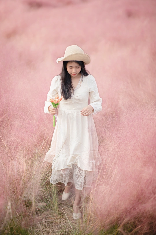 一位穿着白色连衣裙和帽子 手里拿着一朵花站在粉红色草地上 young woman