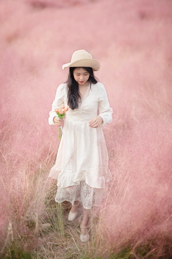 一位穿着白色连衣裙和帽子 手里拿着一朵花站在粉红色草地上 young woman