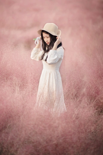 一位穿着白色连衣裙和帽子站在粉红色草地的年轻女子