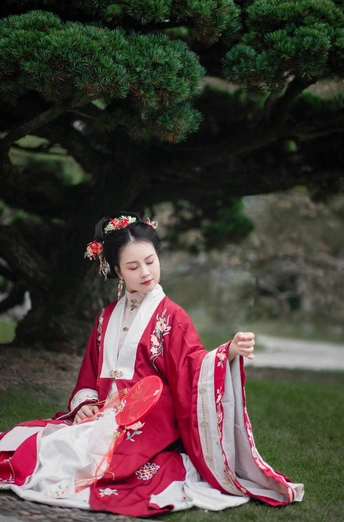 一位穿着传统服装的年轻女子坐在一棵树前的草地上。