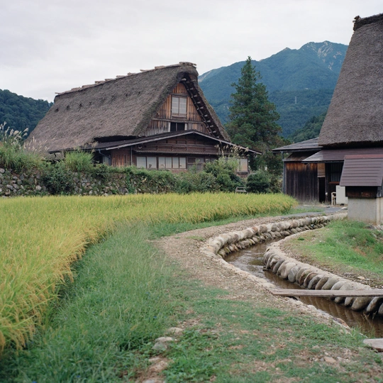 前景有一条流水 背景有一座房子 周围是草地和山。