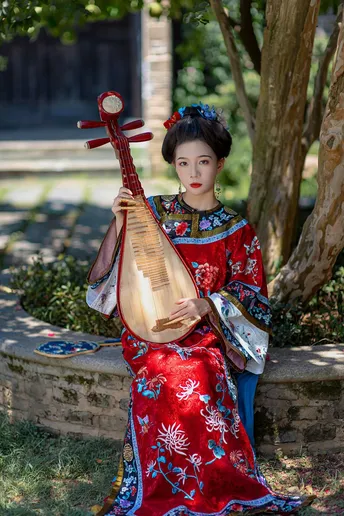 一位穿着传统服装的年轻女子坐在树下 穿着红色裙子 同时拿着乐器。