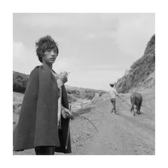 一张黑白照片 一位年轻女子穿着外套走在一条泥土路上 一匹马和一头牛站在路上。