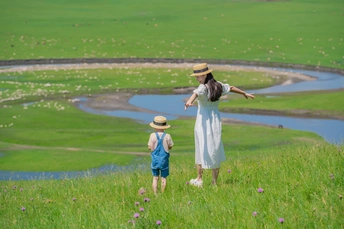 一个戴着帽子的年轻女人和一个小女孩站在小山上 看着绿色草地上的池塘