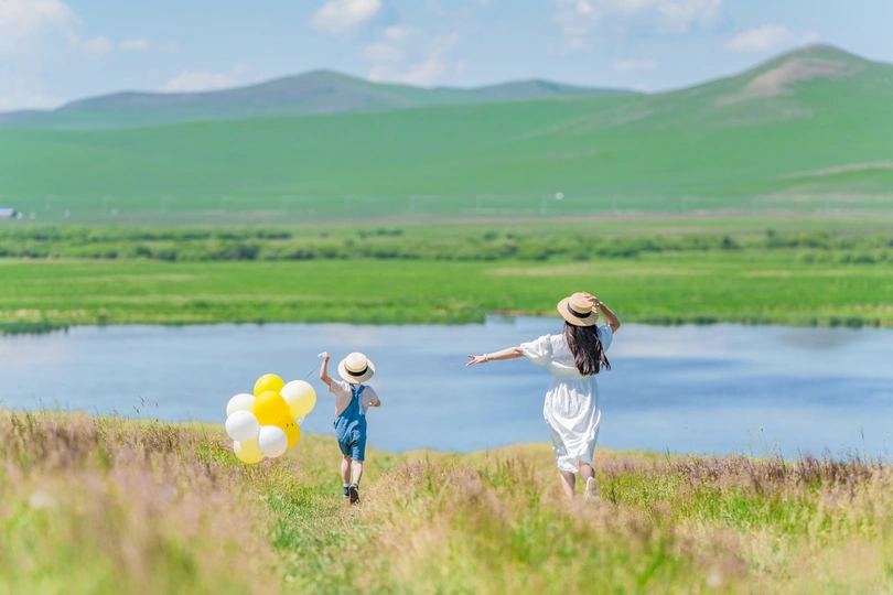 一个女孩拿着气球在草地上奔跑 一个孩子在背景中奔跑 穿过一片绿色的草地 草地上有绿色的山丘和一条河