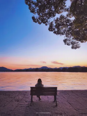 夕阳下坐在长凳上 树下俯瞰水面的一个人。