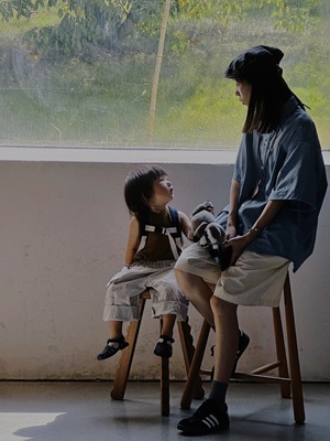 一位妇女坐在小椅子上 正在窗口看着一个坐在小板凳上的小女孩。