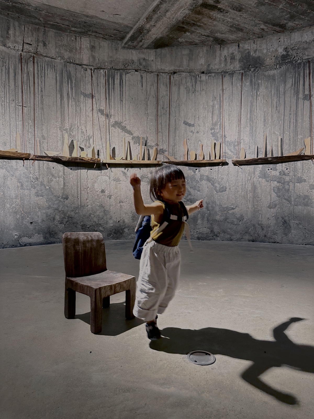 一个小男孩在一个带有混凝土墙壁和一个小型木凳的房间里玩着一把木椅子。