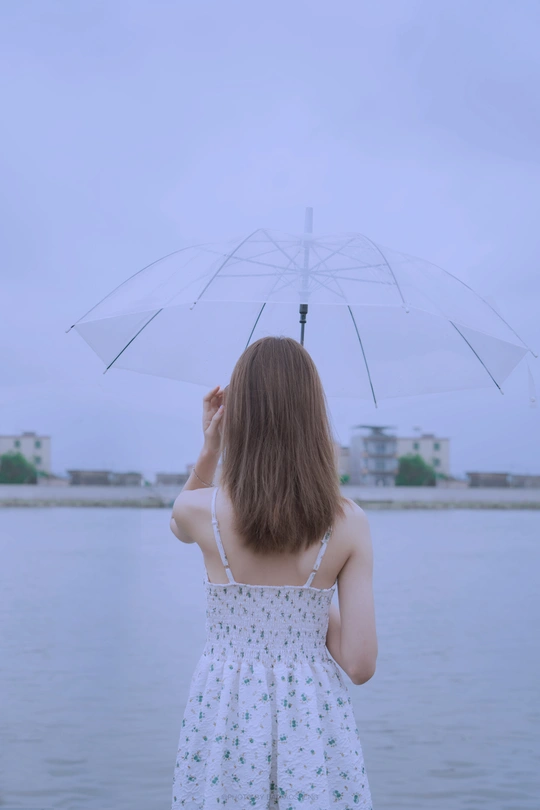一位穿着白色连衣裙的年轻女子手持雨伞 眺望着一片水域。