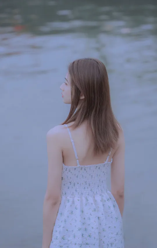 一位穿着白色连衣裙的年轻女子沿着海滩走向水边 注视着水面。